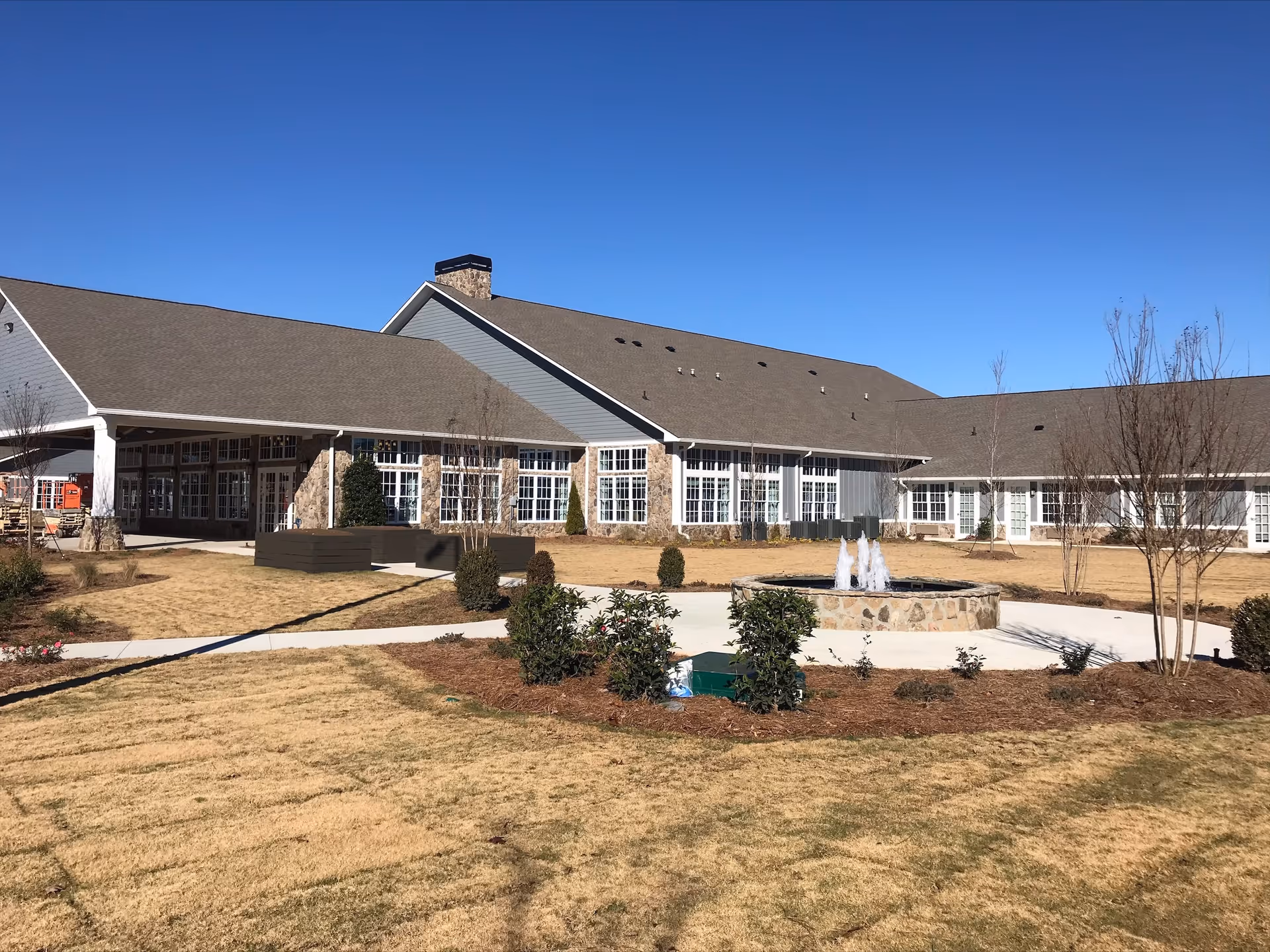 Exterior view of Manor Lake Assisted Living & Memory Care - Hoschton building with a large lawn, landscaped bushes, a stone fountain with water jets, and clear blue sky.