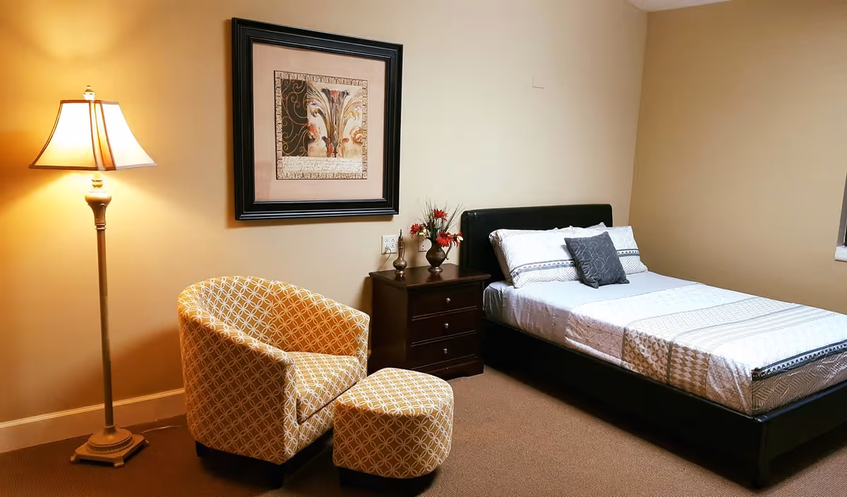 A cozy bedroom with a bed featuring white and patterned bedding, a dark wooden nightstand with decorative vases and flowers, a patterned armchair with a matching ottoman, a standing lamp, and a framed artwork on the wall.