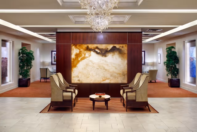 A polished lobby seating area with four armchairs around a low round table, a large illuminated stone art panel, chandeliers, and potted plants.