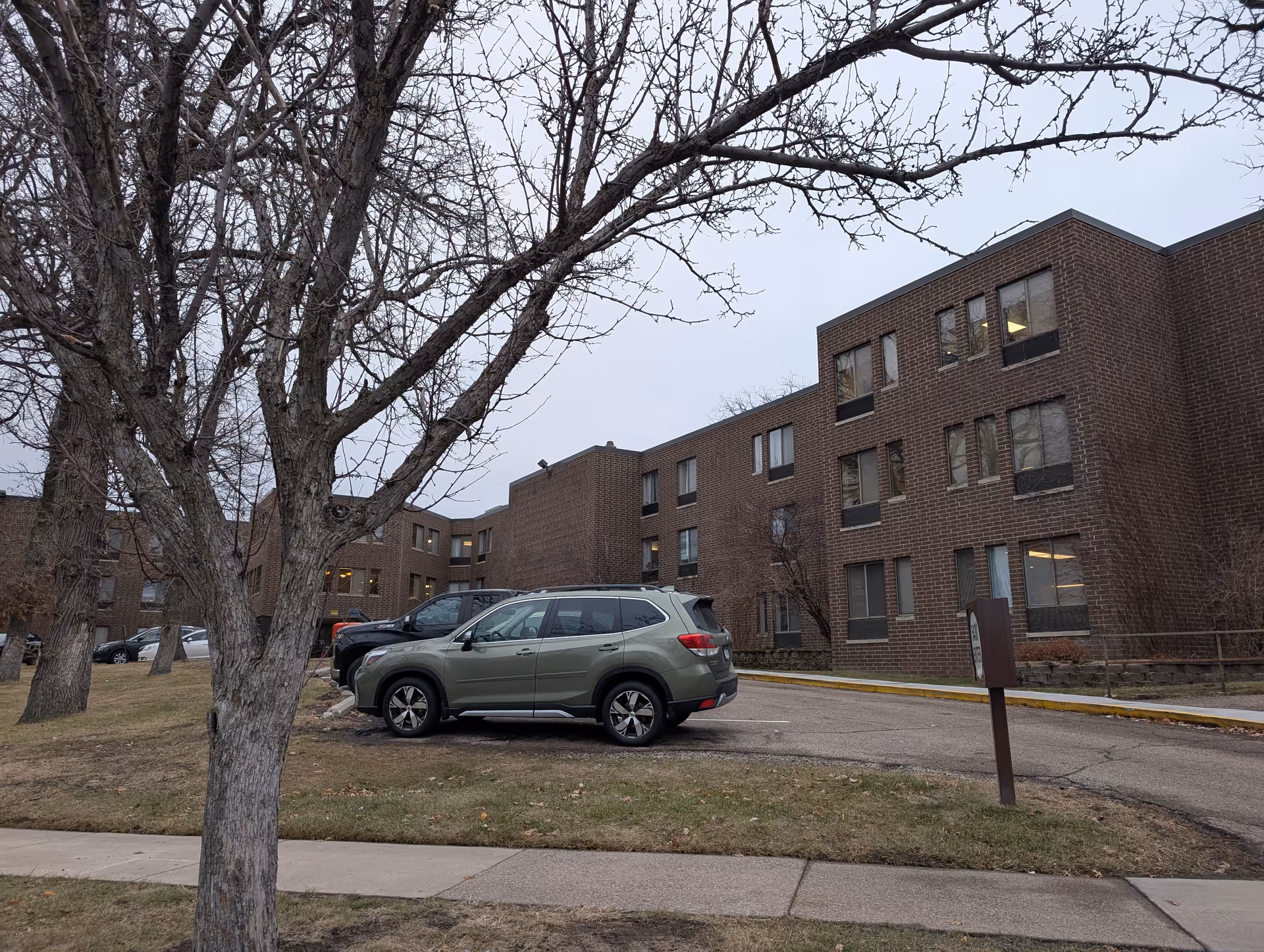 Exterior view of a brick building with multiple windows, a parking area with several parked cars, and leafless trees in the foreground under an overcast sky.