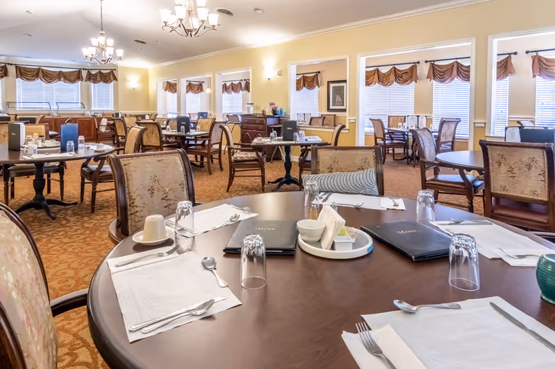 A bright assisted-living dining room with round tables set for meals, upholstered chairs, chandeliers, and windows with valances.