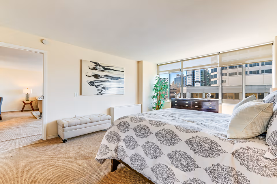 Sunlit bedroom with a large bed featuring patterned bedding, a tufted bench, dresser and floor-to-ceiling windows overlooking the city.