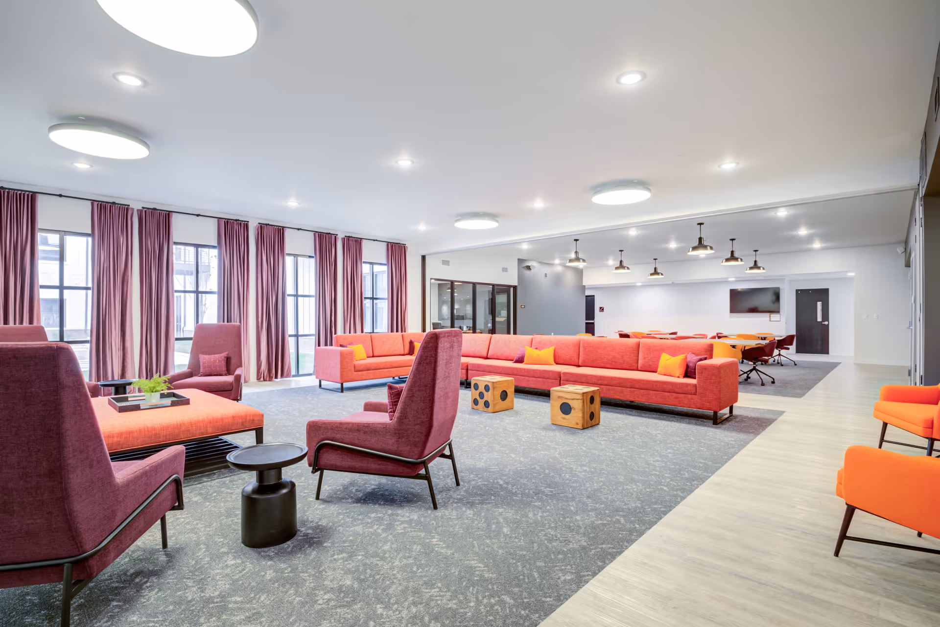 A spacious and modern living room area in a senior living facility with large windows covered by purple curtains. The room features a long red sectional sofa with orange and purple pillows, several purple armchairs, two wooden cube stools, and a small black side table. The floor is a combination of carpet and light wood, and the ceiling has multiple round and pendant lights. In the background, there is a conference or dining area with orange chairs and a wall-mounted TV.