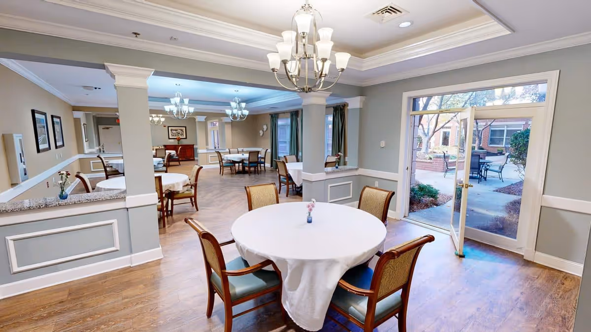 Interior view of a senior living facility dining room with round tables covered in white tablecloths and wooden chairs with cushioned seats. The room has light gray walls with white trim, chandeliers hanging from the ceiling, and large windows and glass doors that open to an outdoor patio area with seating and greenery.