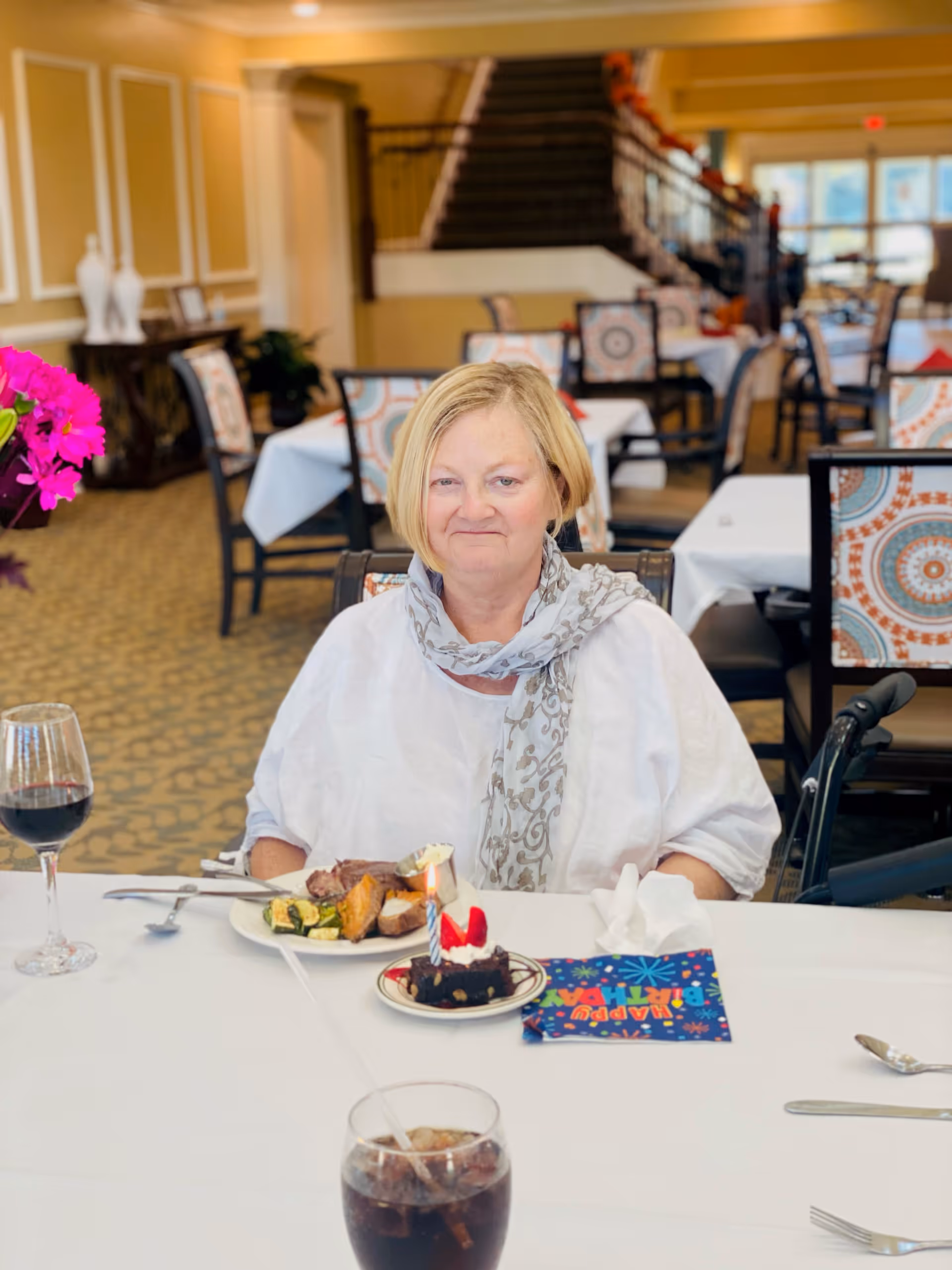 A woman sits at a dining table in a decorated dining room with a plated meal, dessert, and drinks.