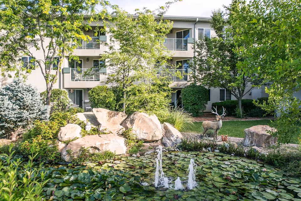 Garden courtyard with a lily-pad pond and small fountain in front of a three-story residential building with balconies, rocks, trees, and a deer statue.