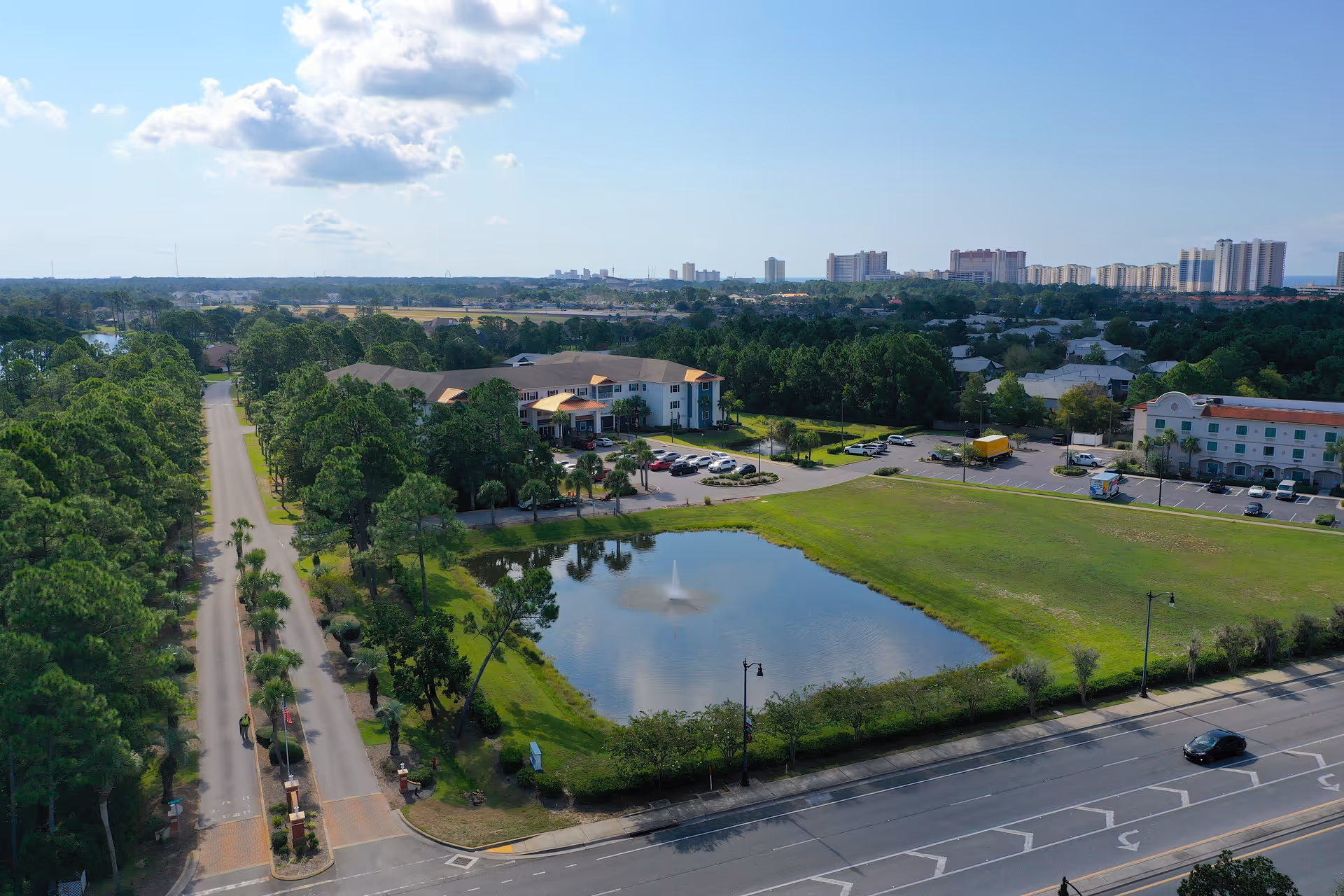 Aerial view of Superior Residences of Panama City Beach showing a large building surrounded by trees, a pond with a fountain, parking lots, and a road with cars. In the background, there are multiple tall buildings and a clear blue sky with some clouds.