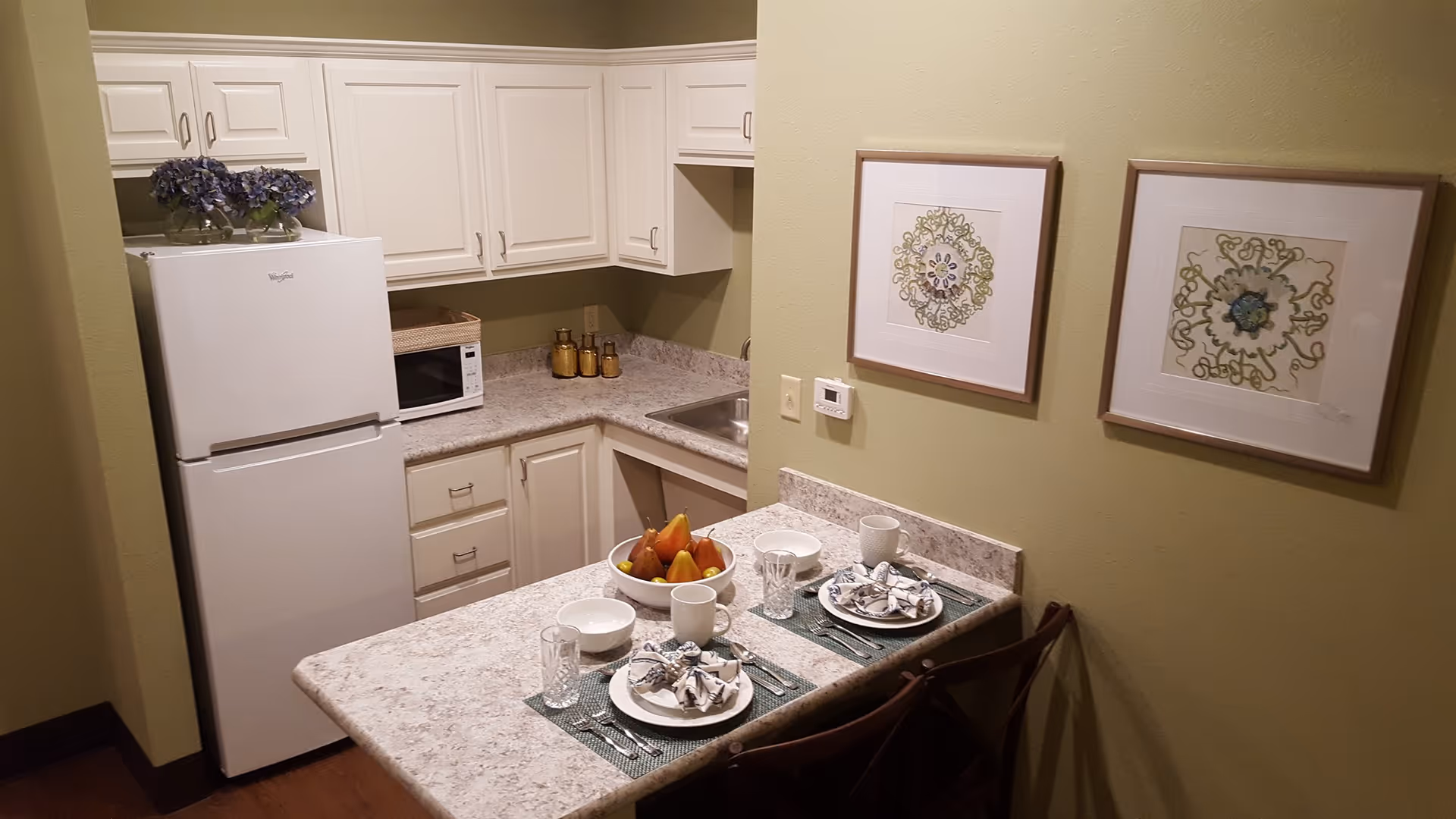Small kitchenette with refrigerator, sink and white cabinets, a breakfast bar set for two with plates and fruit, and framed artwork on a green wall.