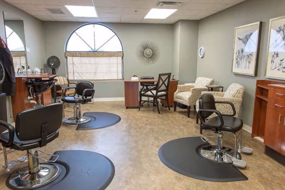 Interior view of a salon area in a senior living facility with three black salon chairs on black mats, a hair washing station, two patterned armchairs, a small table with two chairs, and wall decorations including framed artwork and a sunburst mirror. There is a large arched window with a beige curtain.