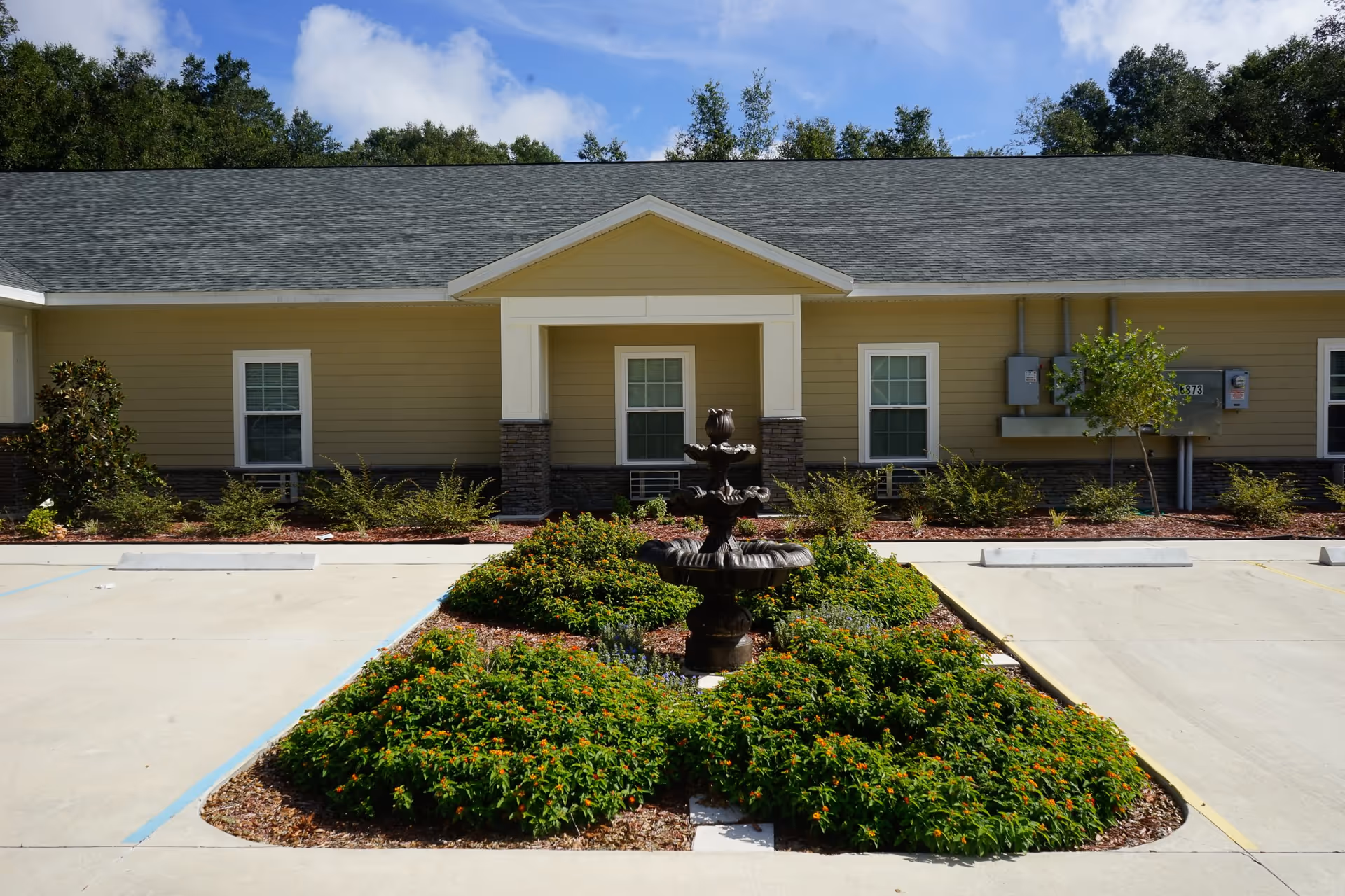 Exterior view of a single-story building with beige siding and a gray shingled roof. In front of the building is a small landscaped area with green bushes and a decorative black fountain. The sky is partly cloudy and there are trees in the background.
