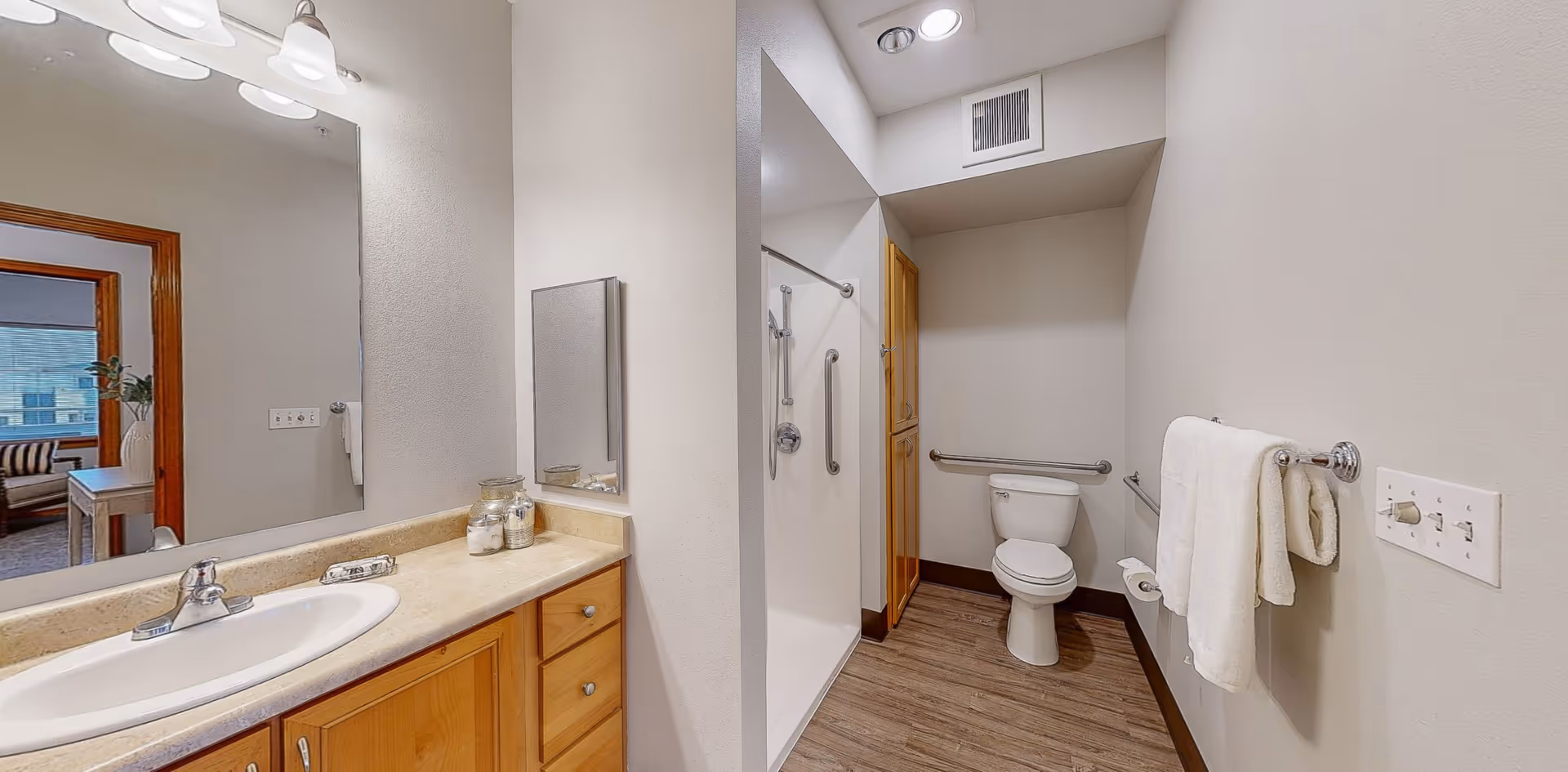 A clean and modern bathroom with a wooden vanity featuring a white sink and countertop. Above the vanity is a large mirror with three light fixtures. To the right, there is a walk-in shower with grab bars and a showerhead. Further back, there is a toilet with grab bars on the walls for accessibility. The bathroom has light-colored walls and wood-look flooring. A towel rack with white towels is mounted on the right wall, and a light switch panel is visible nearby. Through the doorway, part of a living area with a window and furniture is visible.
