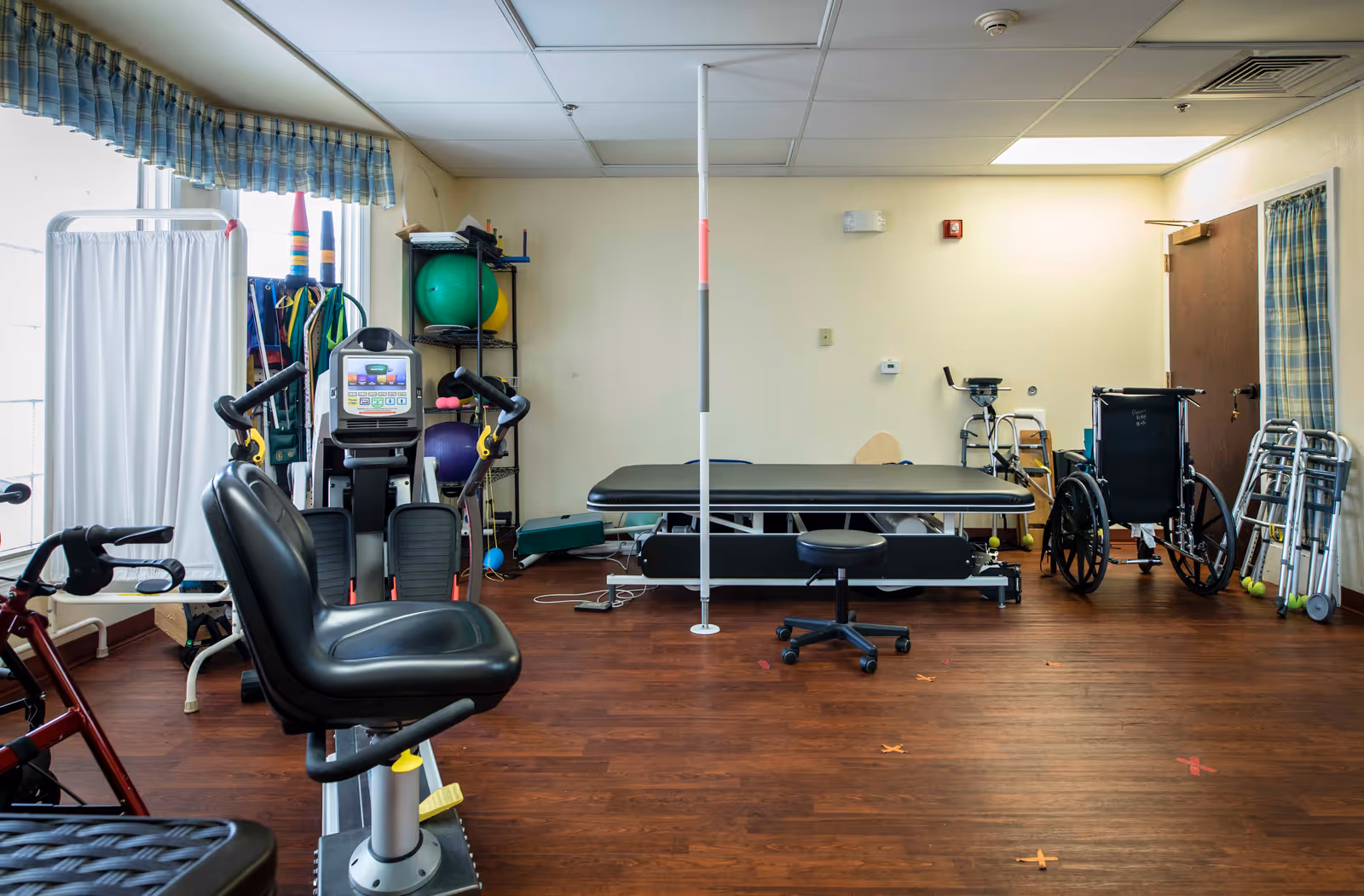 A senior living therapy room with exercise machines, a treatment table, wheelchairs, walkers, and fitness equipment on a wood floor.