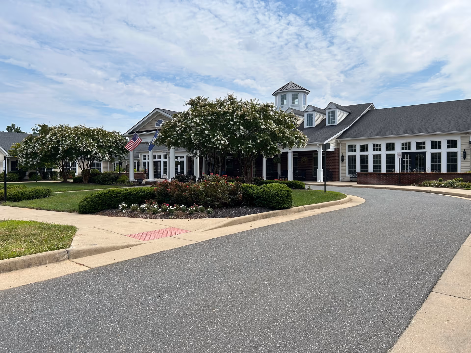 Front driveway and main entrance of a single-story senior living building with landscaped flowerbeds, crepe myrtle trees, an American flag and a small cupola.