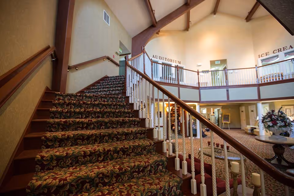 Carpeted staircase with floral pattern leading to an upper level inside a senior living facility. The upper level has white railings and signs indicating an auditorium and an ice cream area. The lower level features a seating area with a round table and floral arrangement, and the space is warmly lit with beige walls and wooden beams on the ceiling.