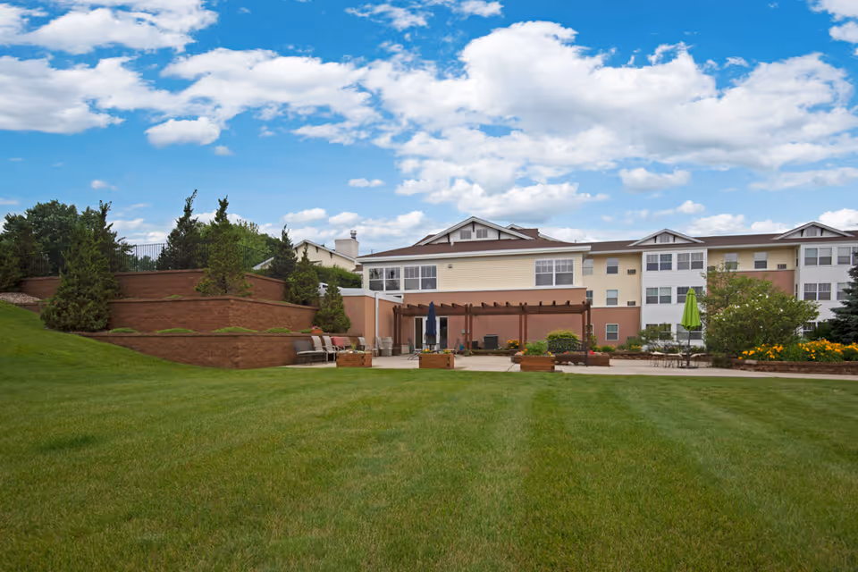 Well-maintained senior living building with a wide green lawn, patio seating and a pergola under a partly cloudy sky.