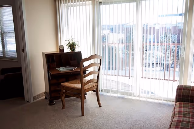 A small wooden desk with a chair in front of large sliding glass doors covered with vertical blinds, allowing natural light to fill the room. There is a small clock and a plant on the desk, and a plaid upholstered chair is partially visible on the right side.