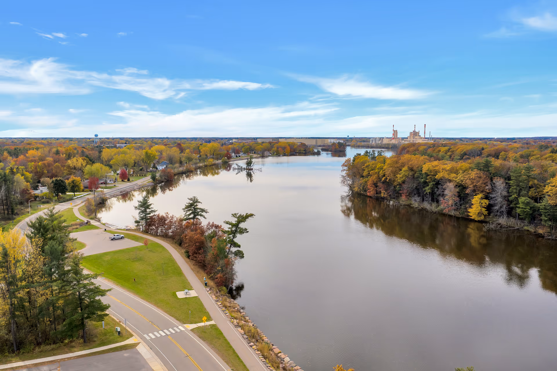 Aerial view of a river surrounded by trees with autumn foliage, a road running alongside the river, and a few parked cars. In the distance, there is an industrial building with smokestacks under a partly cloudy blue sky.