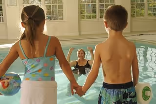Two children holding hands stand at the edge of an indoor swimming pool, looking at an adult woman in the water who is raising her arms. The pool area has large windows letting in natural light.