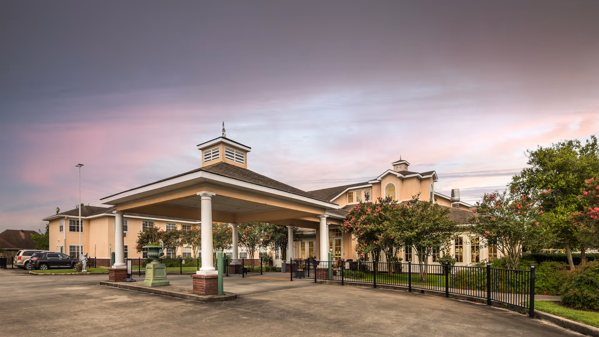 Exterior view of Homestead Assisted Living facility during sunset with a covered entrance supported by white columns, surrounded by trees and parked cars.