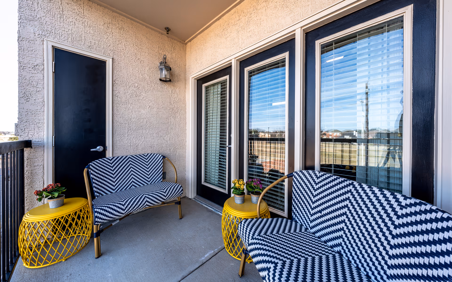 Outdoor balcony area with two black and white patterned cushioned benches and two yellow metal side tables with small potted plants. The balcony has a textured beige wall, a black door, and large windows with blinds reflecting the outdoor view.