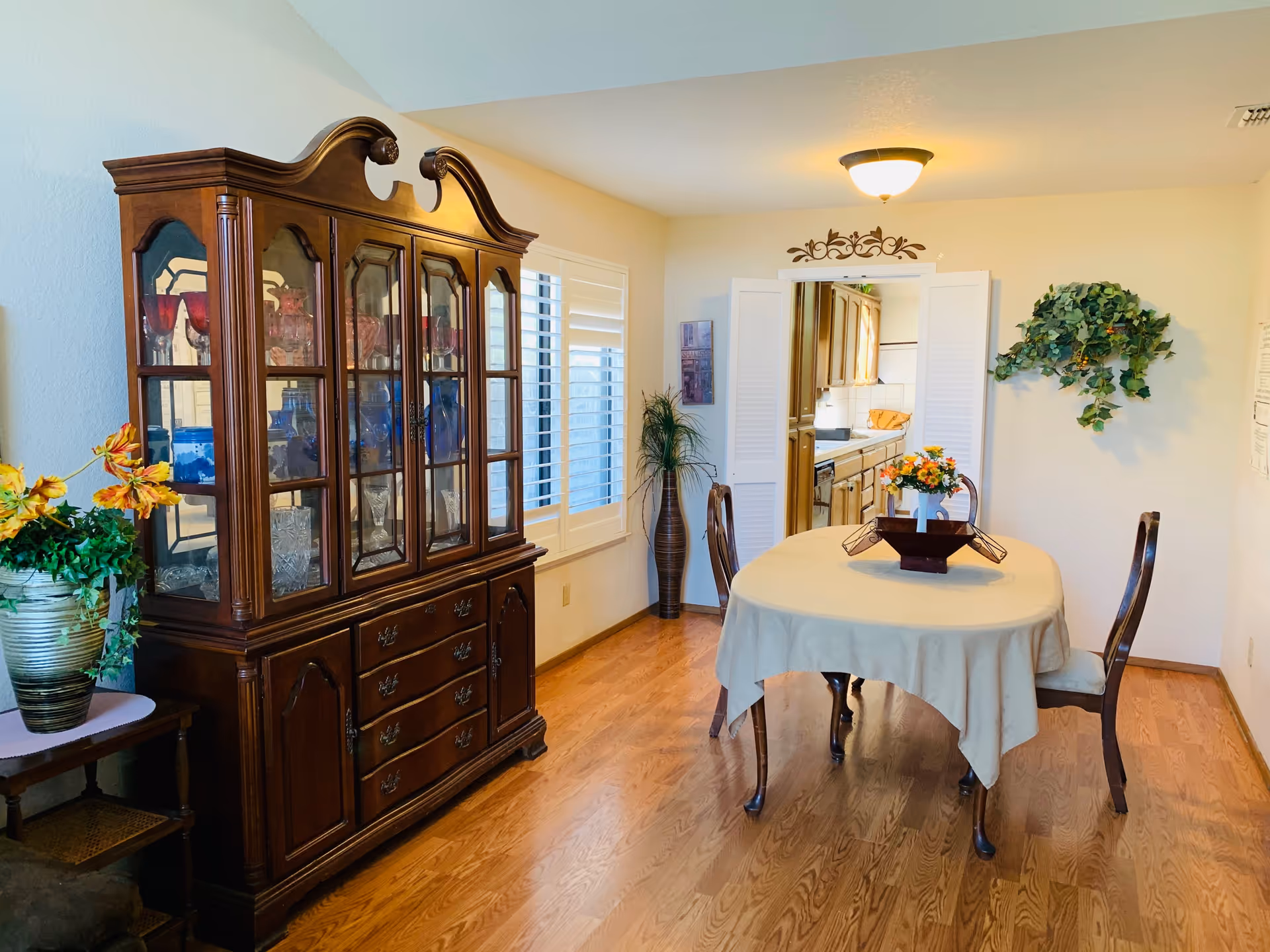 A dining room with a wooden floor, a table covered with a beige tablecloth, and two wooden chairs. On the table is a decorative centerpiece with flowers. To the left is a large wooden china cabinet displaying glassware and decorative items. A window with white shutters is on the left wall. The room leads into a kitchen through an open doorway with white folding doors. There is a wall decoration above the doorway and a green leafy plant mounted on the right wall.