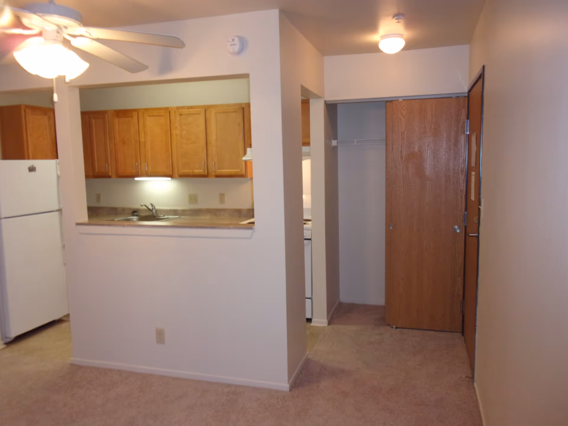 Interior view of a senior living facility apartment showing a small kitchen with wooden cabinets, a white refrigerator, and a sink. There is a ceiling fan with a light on the left side and a hallway with a wooden door and a closet on the right side. The walls are painted white and the floor is carpeted.