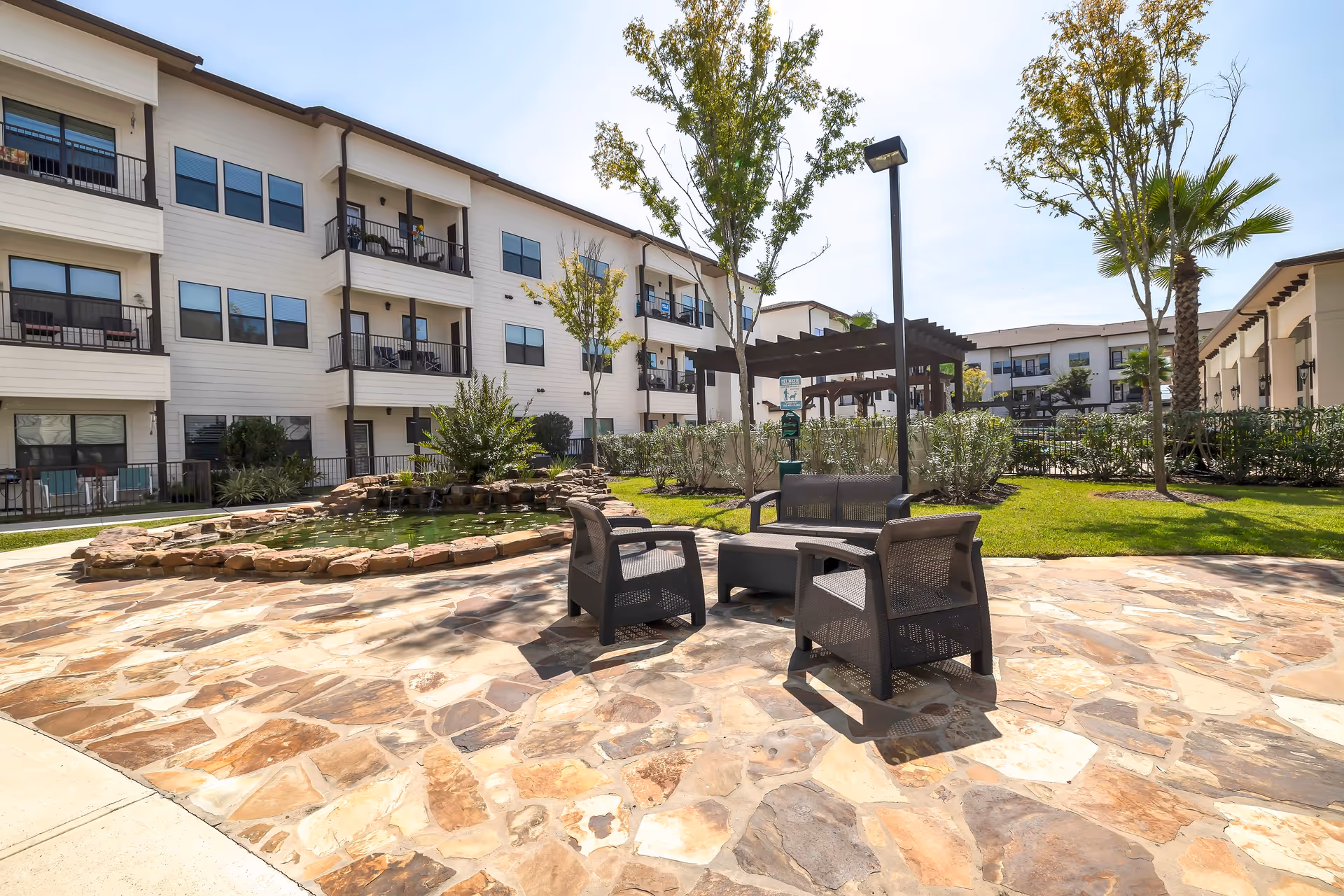 Outdoor courtyard area at Solea Copperfield featuring a stone-paved patio with black outdoor chairs and a small table. There is a pond with rocks around it, green grass, trees, and a pergola structure in the background. The courtyard is surrounded by a three-story residential building with balconies and windows.