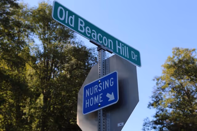 Street signs at a road intersection showing 'Old Beacon Hill Dr' and a blue sign indicating the direction to a nursing home, with trees and a clear blue sky in the background.