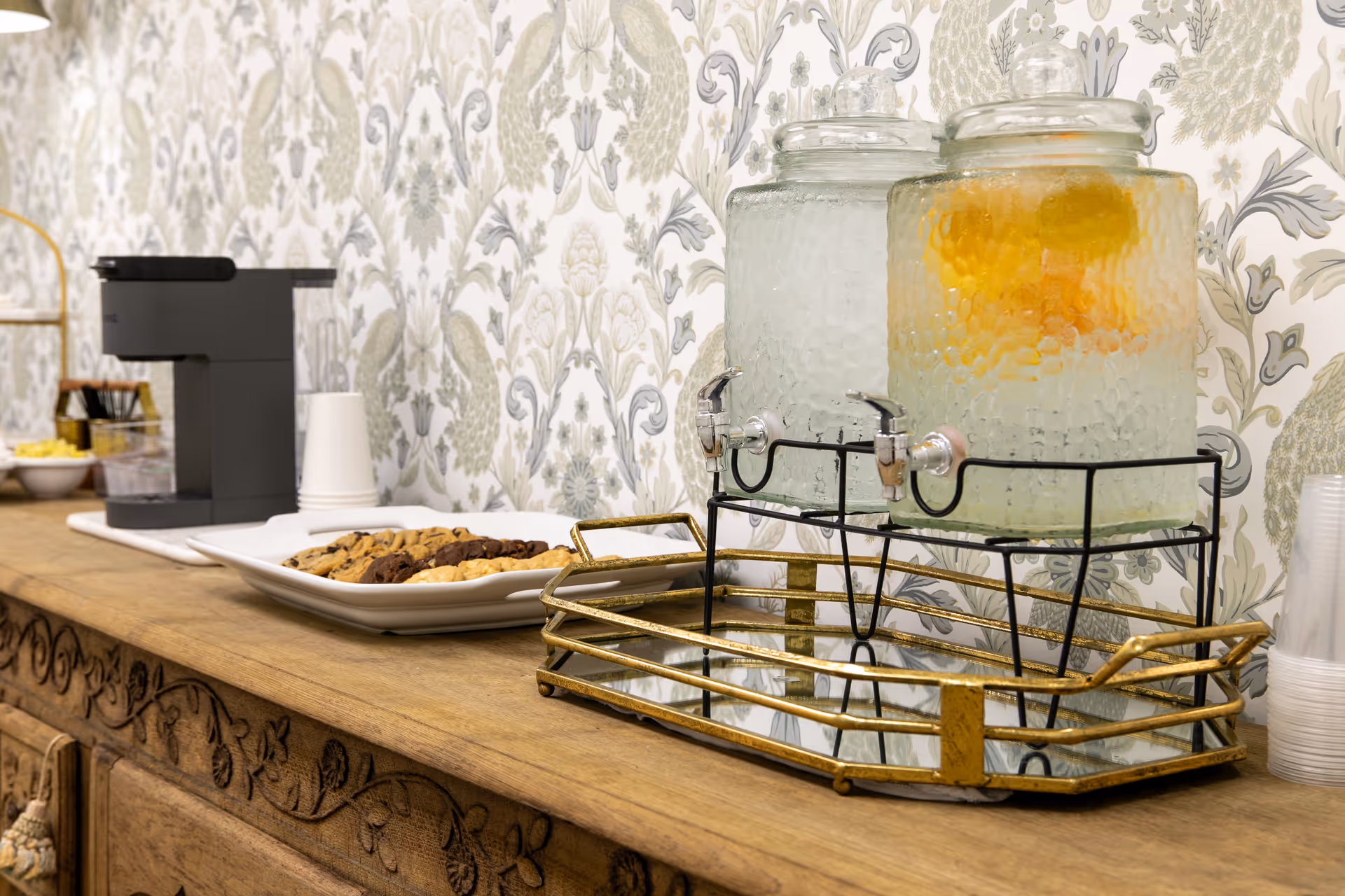 A wooden sideboard with two glass beverage dispensers, one filled with water and the other with a citrus-infused drink, a plate of assorted cookies, a coffee machine, disposable cups, and a decorative floral wallpaper background.