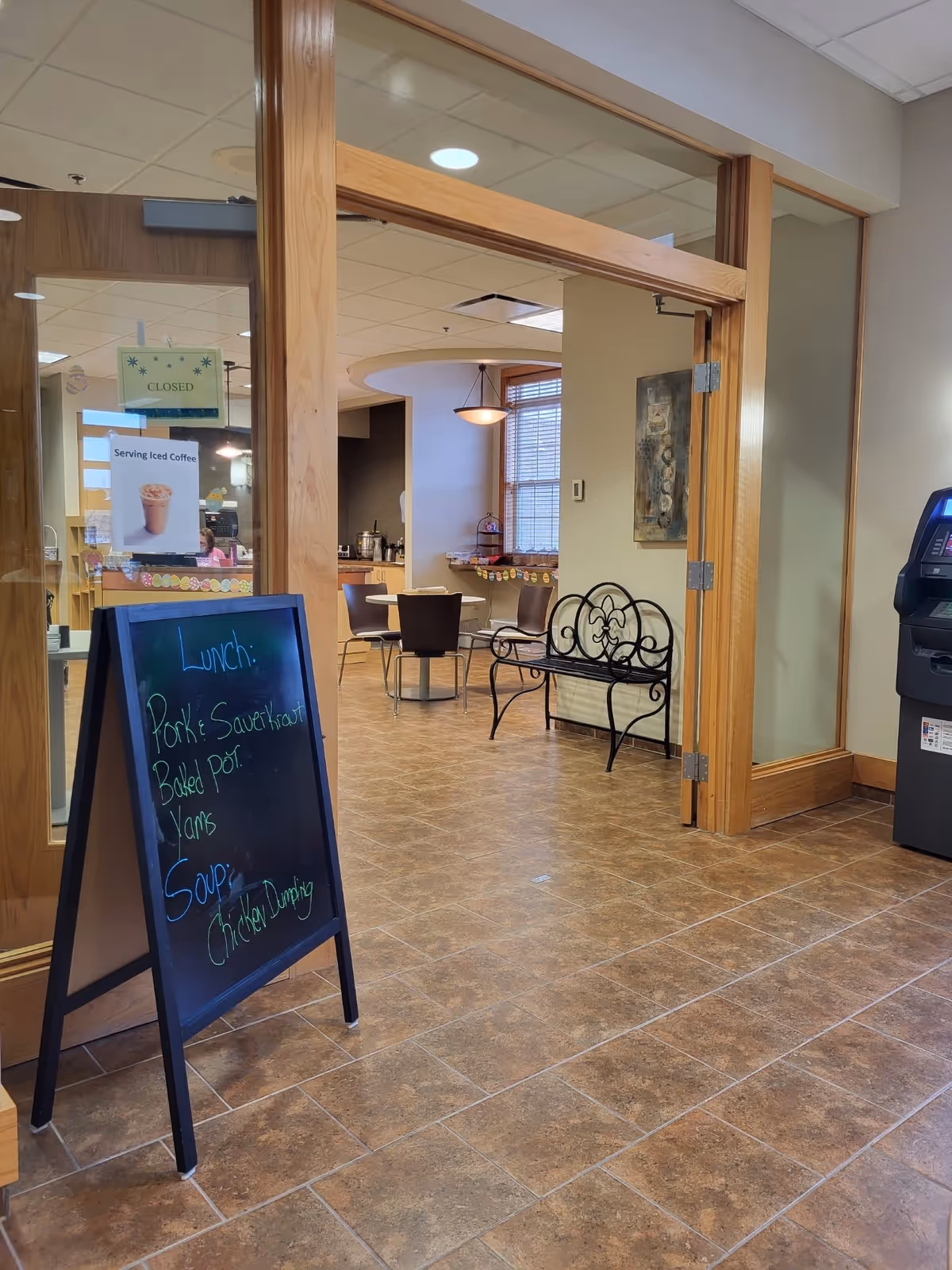 Interior view of a senior living facility dining area with a blackboard sign listing lunch menu items including pork and sauerkraut, baked potato, yams, and chicken dumpling soup. The area has tiled flooring, a wooden-framed glass door, a metal bench, and a table with chairs near a window.