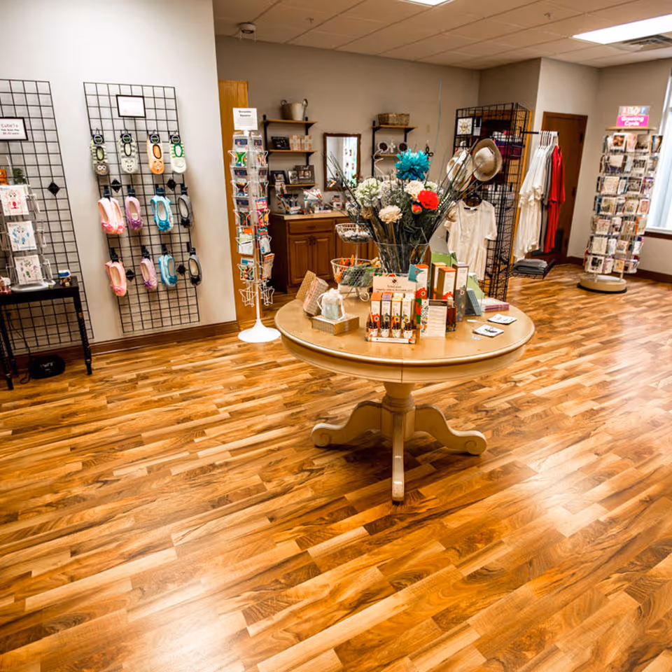 Interior view of a small retail or gift shop area with wooden flooring, a round table in the center displaying various products and a vase with colorful flowers. Shelves and racks around the room hold clothing, slippers, greeting cards, and other items. The space is well-lit with a window on the right side.