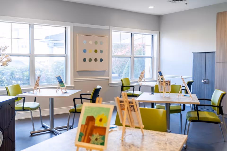 A bright room with several tables and green chairs arranged for an art activity. Each table has small wooden easels holding paintings or drawings. Large windows let in natural light, and a colorful abstract painting hangs on the wall.