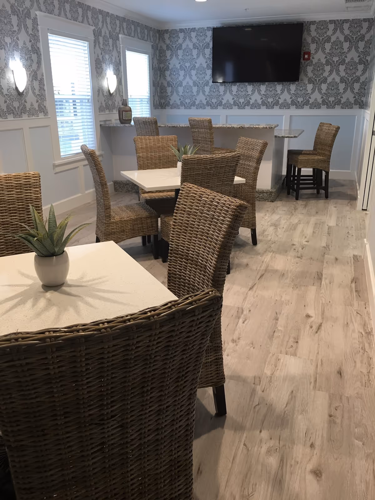 A dining area with several square white tables and wicker chairs. The room has light wood flooring, patterned wallpaper on the upper half of the walls, and white wainscoting on the lower half. There are two windows with blinds on the left wall, a flat-screen TV mounted on the far wall, and a countertop with bar stools in the background. Small potted plants are placed on the tables.