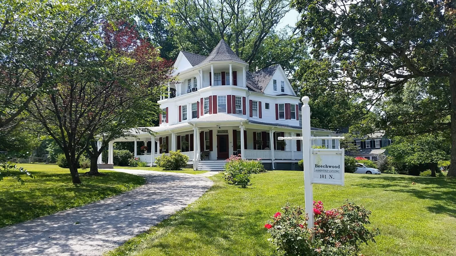 A large white multi-story house with red shutters and a wraparound porch surrounded by green trees and grass on a sunny day. A curved driveway leads up to the house. In the foreground, there is a white signpost with a sign that reads 'Beechwood Assisted Living 101 N.'