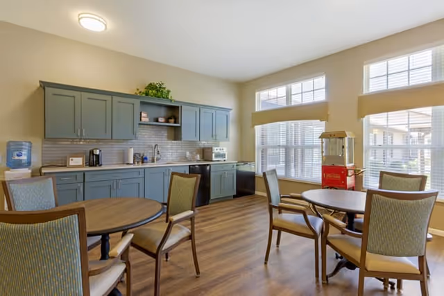 Bright communal dining area with round tables and chairs, a kitchenette with blue-green cabinets, and large windows.