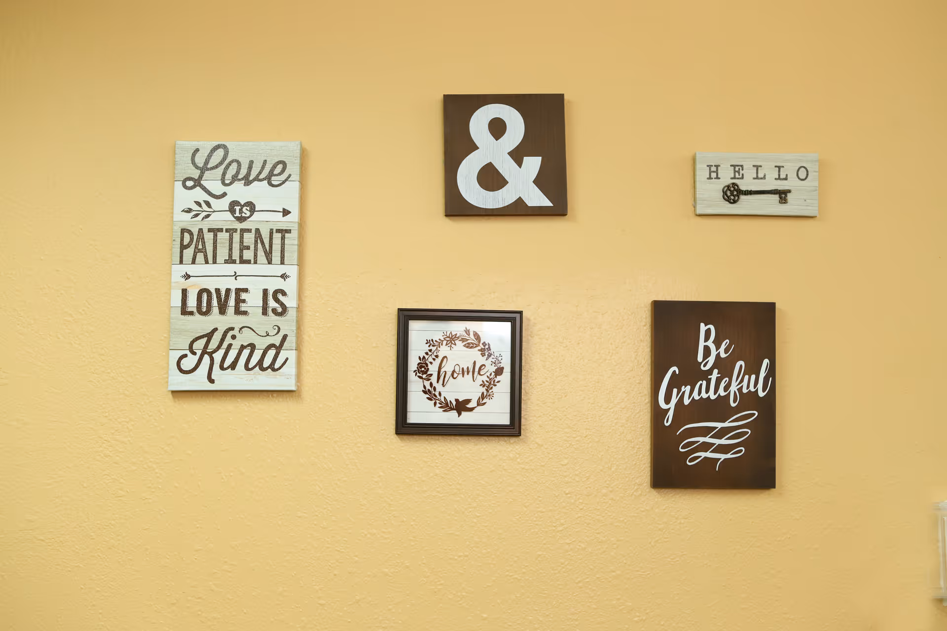 A beige wall with five decorative wooden plaques. The plaques have various inscriptions including 'Love is Patient Love is Kind', a large ampersand symbol, 'HELLO' with a key illustration, 'home' surrounded by a floral wreath, and 'Be Grateful'.