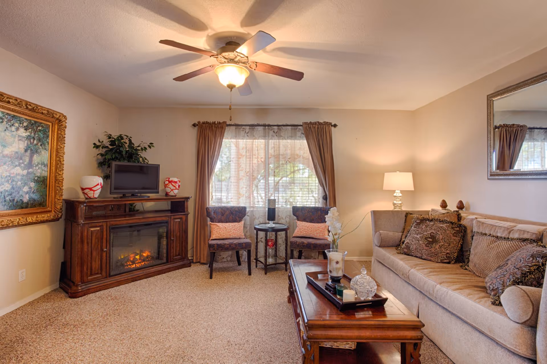 A cozy living room with a beige sofa adorned with patterned cushions, a wooden coffee table with decorative items, two upholstered chairs with pink pillows near a window with sheer and brown curtains, a wooden cabinet with an electric fireplace and a small TV on top, a large framed painting on the left wall, a ceiling fan with a light, and a table lamp on a side table.