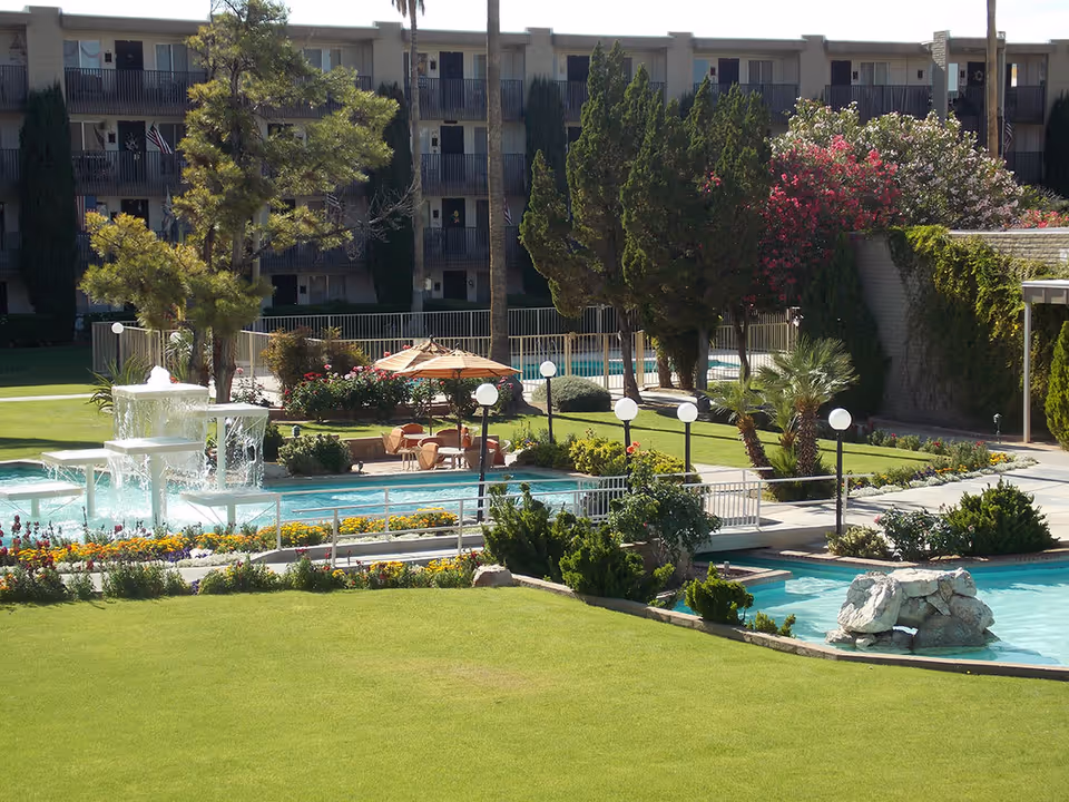Outdoor view of a senior living facility with a well-maintained lawn, a multi-tiered water fountain, a small pool with rock features, and several trees and flowering bushes. There are patio tables with umbrellas and chairs near the pool area, and a multi-story building with balconies in the background.