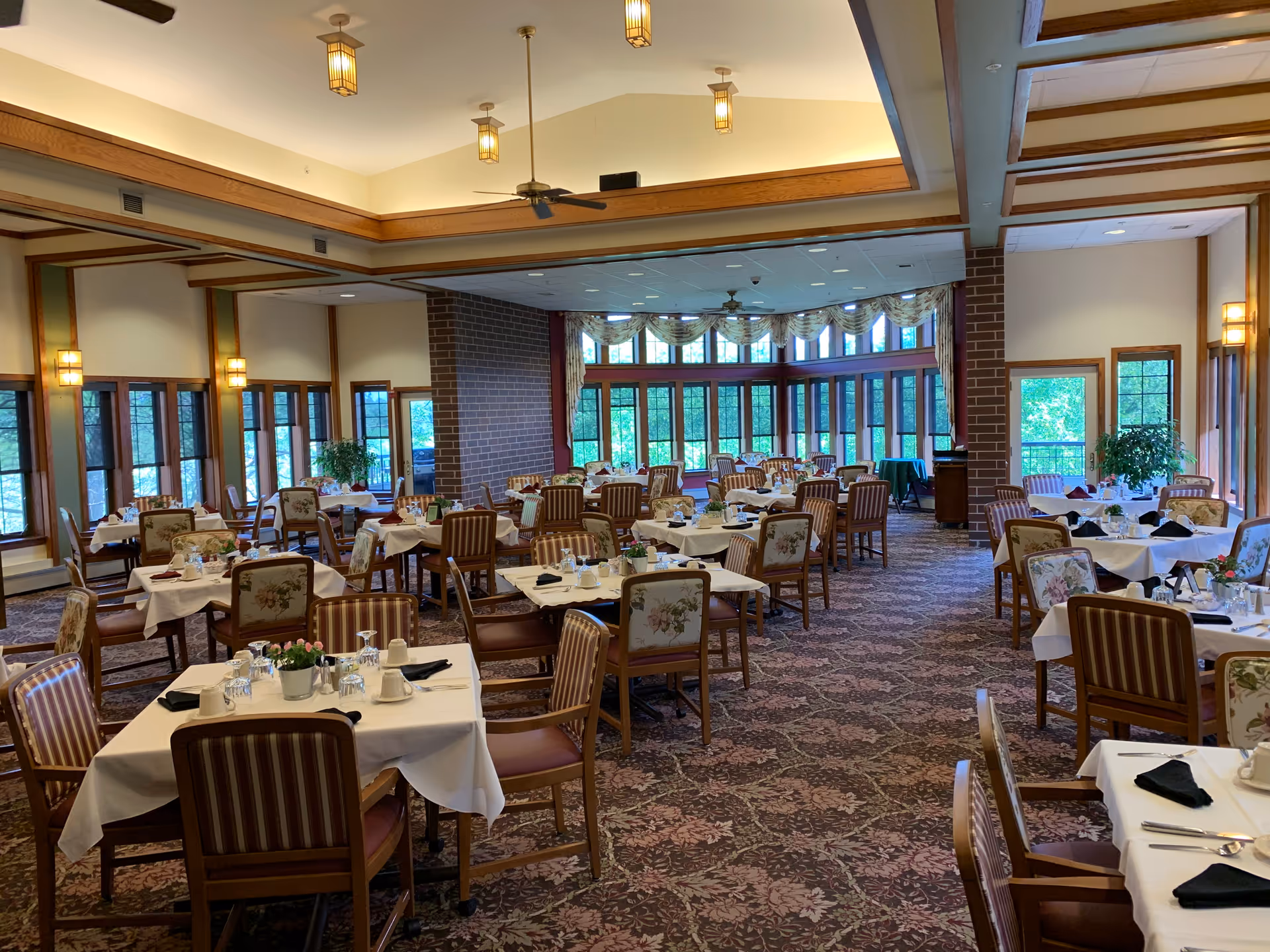 A spacious dining room with multiple tables covered in white tablecloths, each set with cups, glasses, and black napkins. The room features large windows with floral curtains, patterned carpet, and wooden chairs with floral and striped upholstery. Ceiling fans and pendant lights hang from the ceiling, and there are plants near the windows.