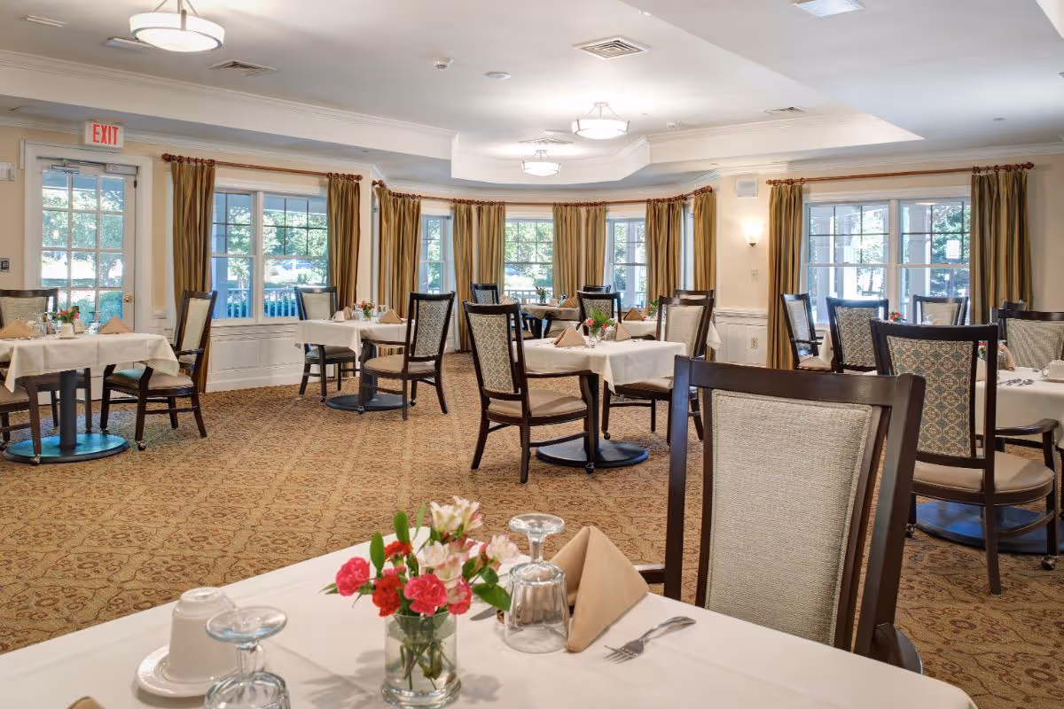 A well-lit dining room with several tables covered in white tablecloths, each set with napkins, glasses, and small flower arrangements. The room has large windows with gold curtains, patterned carpet, and wooden chairs with cushioned seats and backs.