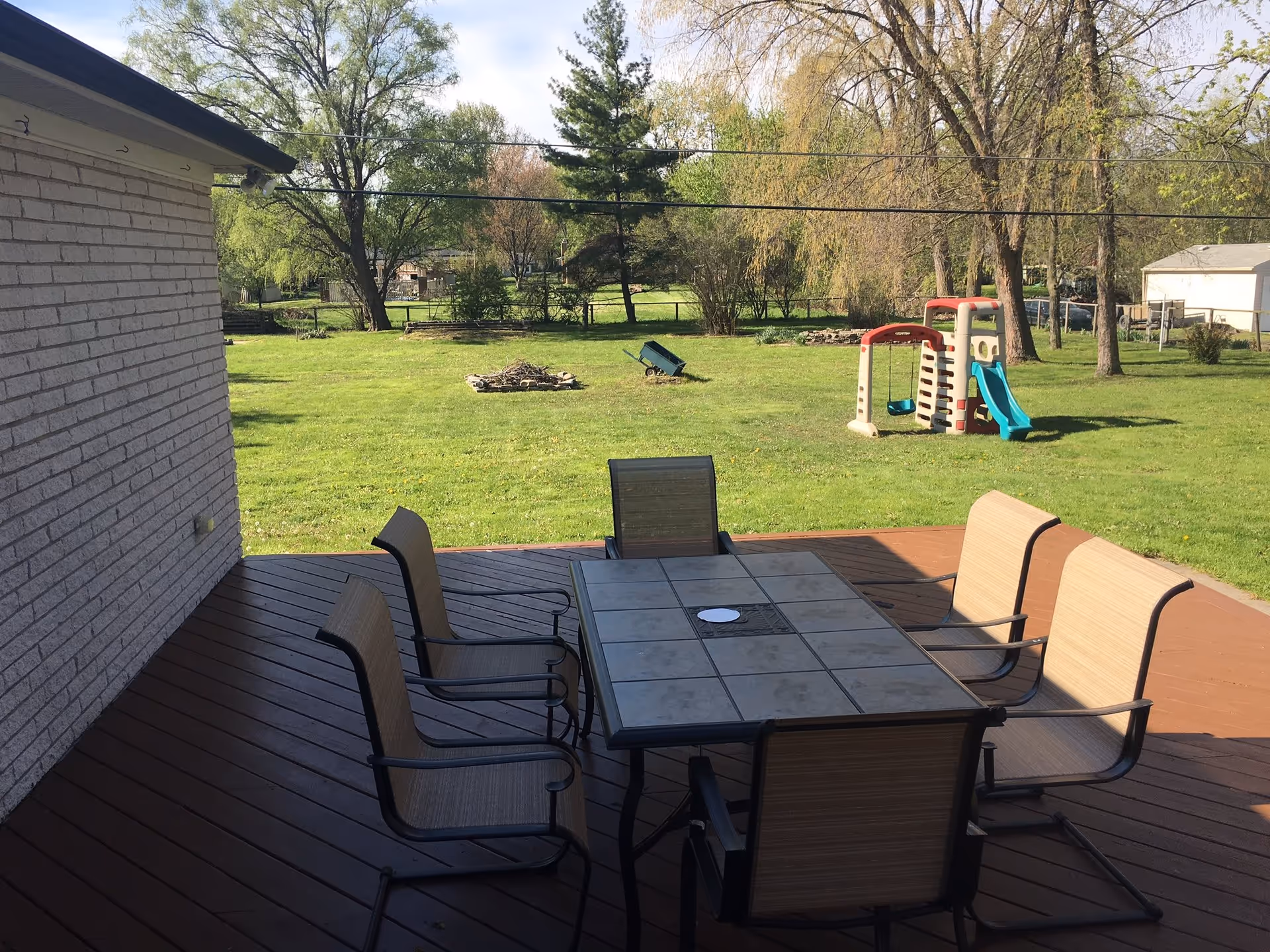 Outdoor wooden deck with a rectangular table and six chairs. Beyond the deck is a grassy yard with trees, a small playground set with a slide and swing, and a fire pit area.