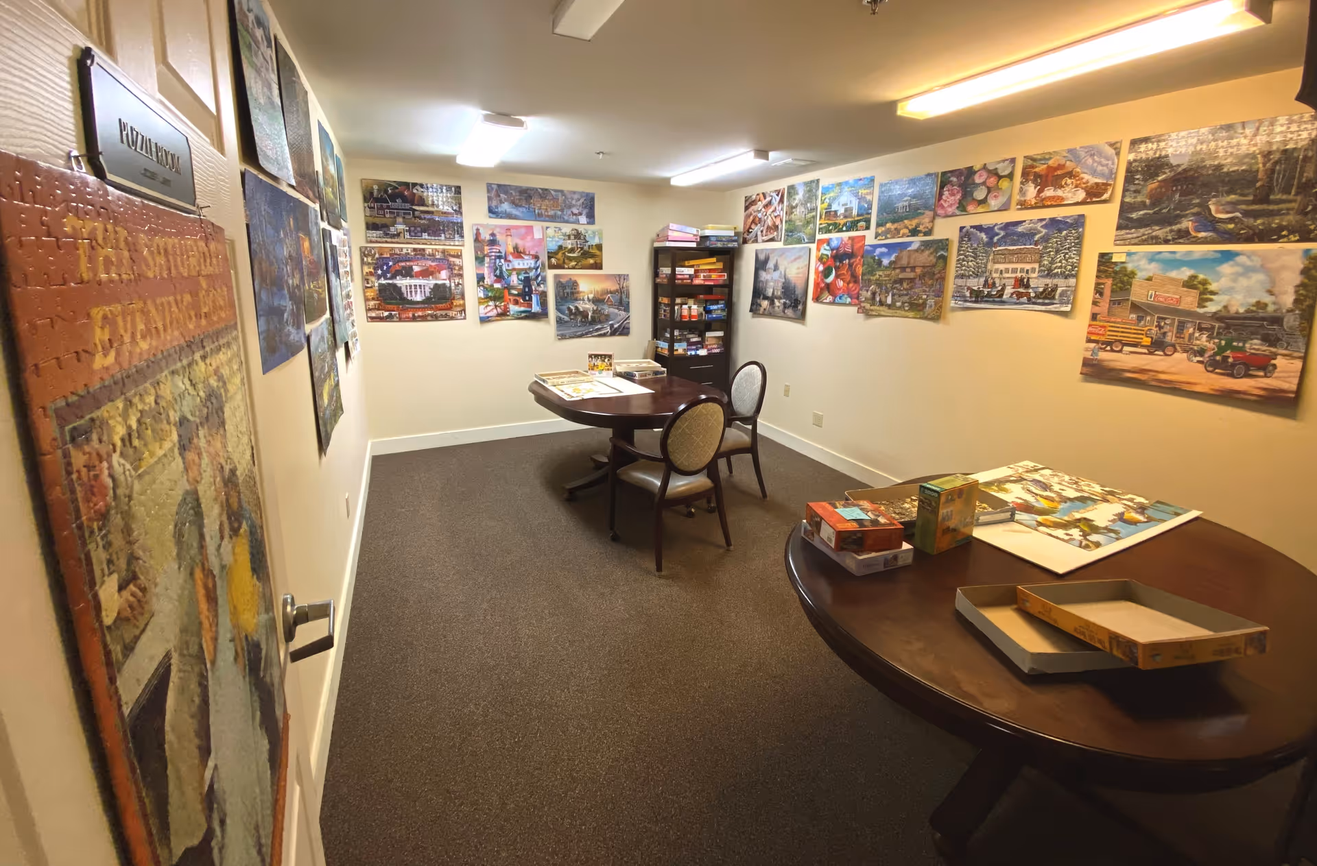 A puzzle room with two round tables and chairs, walls decorated with completed puzzles and puzzle boxes on the tables and a shelf filled with board games and puzzles.