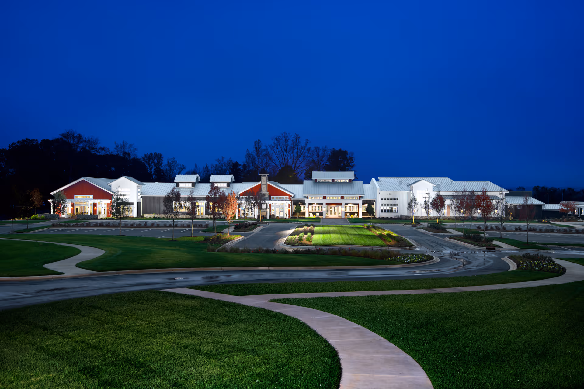 Nighttime exterior view of a large, well-lit senior living facility building with a modern farmhouse design, surrounded by landscaped lawns and curved sidewalks.