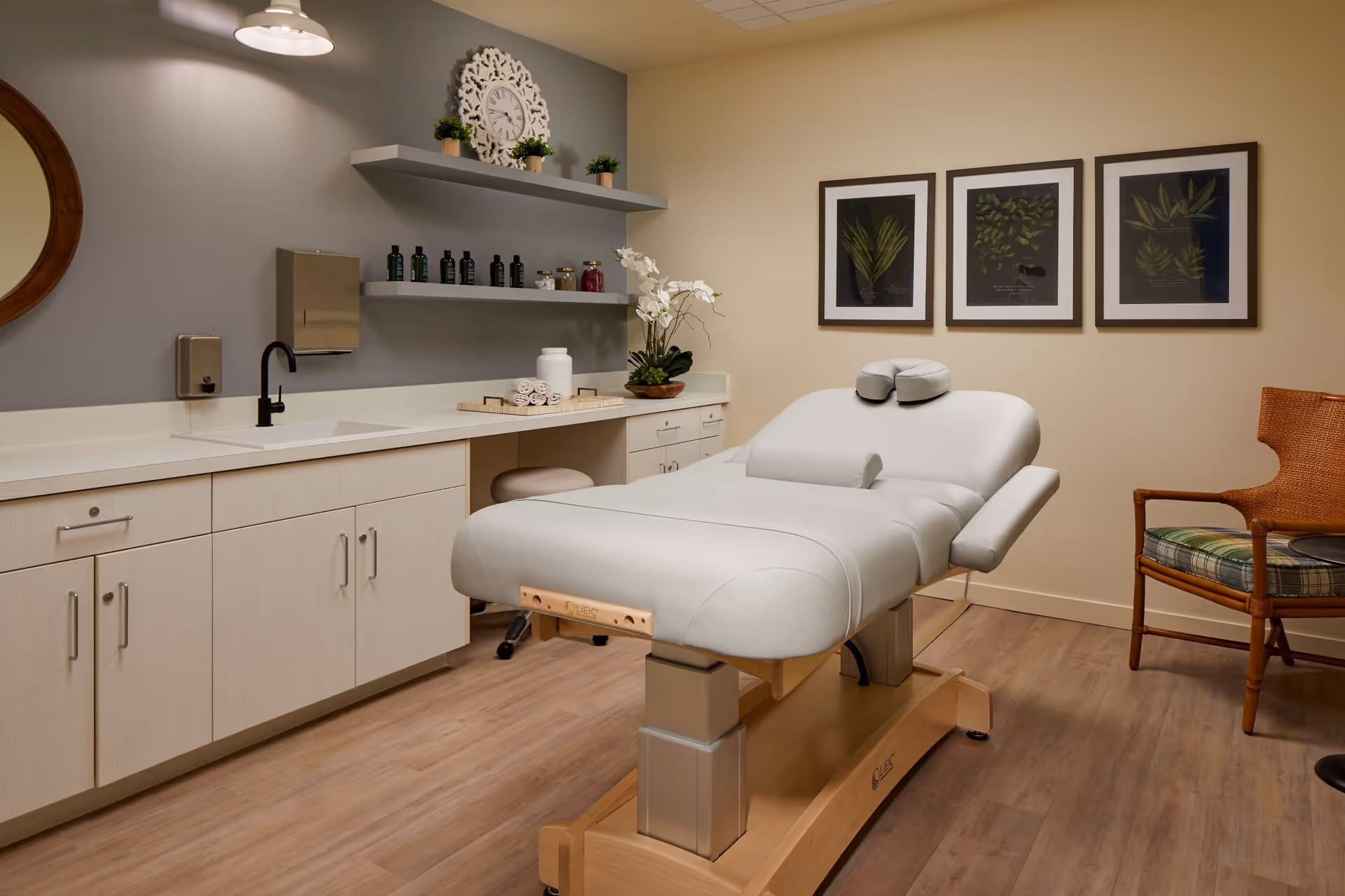 A clean and well-lit therapy or massage room featuring a white adjustable treatment table in the center. The room has light wood flooring, beige walls, and a gray accent wall with two floating shelves holding bottles and decorative items. There is a round mirror on the left wall, three framed botanical prints on the right wall, a wooden chair with a plaid cushion, and a small black side table.