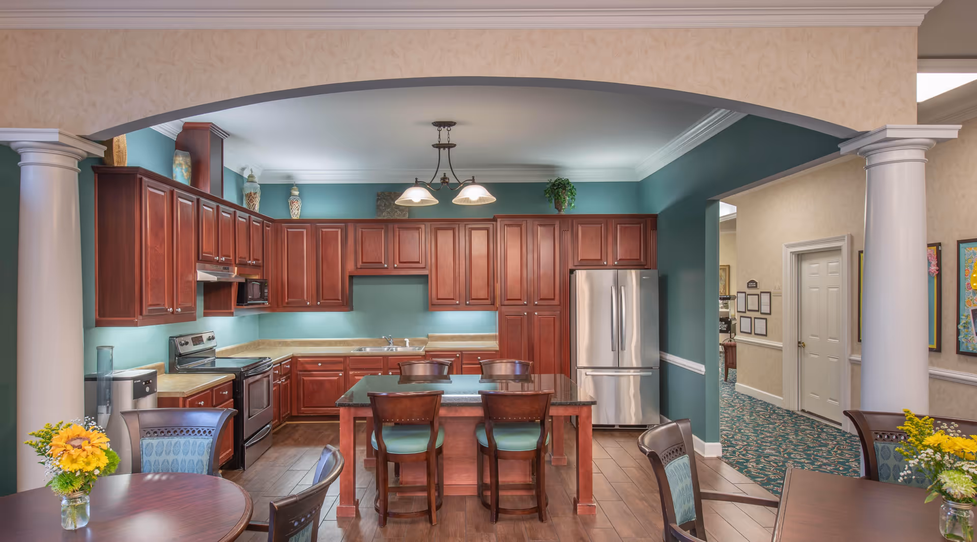A spacious kitchen area with wooden cabinets, stainless steel appliances including a refrigerator and stove, and a central island with four chairs. The kitchen is painted in teal and beige tones with decorative vases on top of the cabinets. In the foreground, there are round dining tables with chairs and small flower arrangements. The room has white columns and an archway leading to a hallway with framed artwork on the walls.