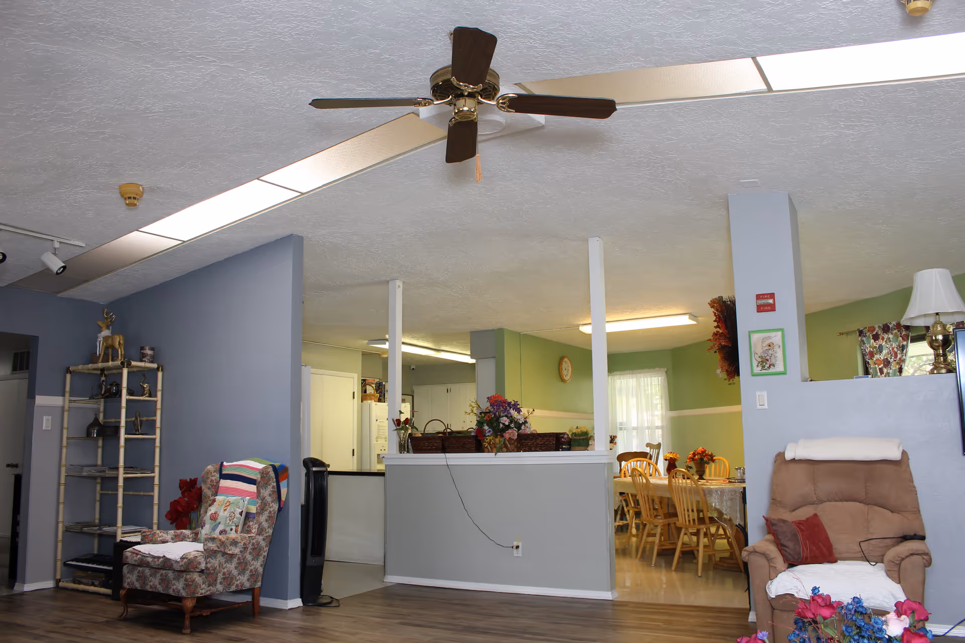 Interior view of a residential care home showing a living room area with a floral armchair and a beige recliner, a ceiling fan, and a dining area with wooden chairs and a table in the background. The walls are painted in shades of blue and green, and there are decorative flowers and a lamp visible.