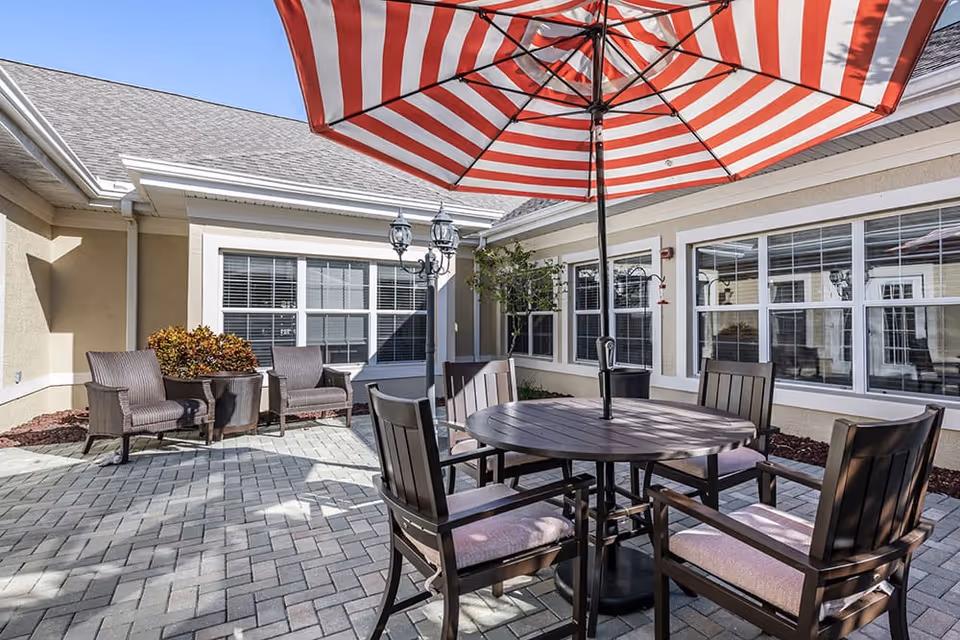 Outdoor patio area with a round wooden table and four cushioned chairs under a red and white striped umbrella. Two additional cushioned chairs and a potted plant are placed against the beige building with multiple windows. The patio is paved with gray bricks and the sky is clear and blue.