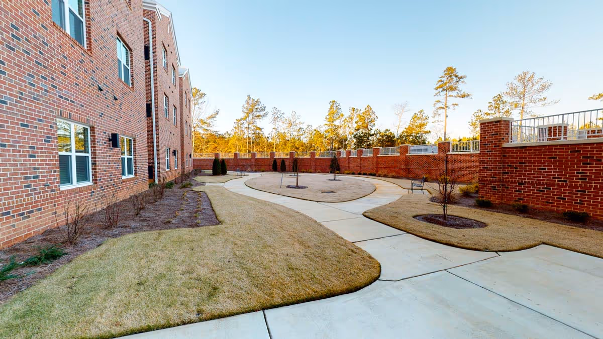 Brick residential building beside a paved winding courtyard path with small landscaped islands and a surrounding brick wall under a clear sky.