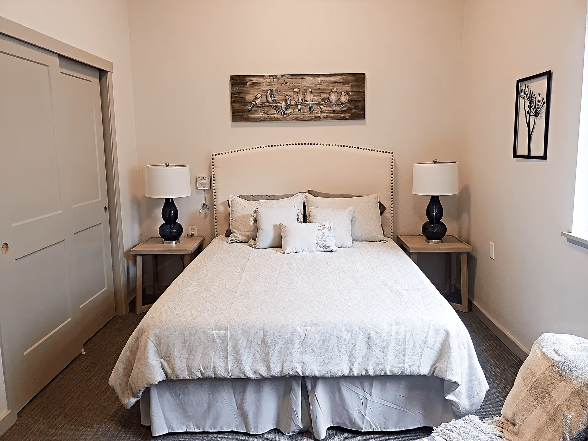 A neatly made bed with a beige upholstered headboard and multiple pillows in a softly lit bedroom. There are two matching bedside tables with dark blue lamps on each side of the bed. Above the bed hangs a wooden artwork featuring birds on a wire. To the left, there is a sliding closet door, and to the right, a window with natural light coming in. A framed plant artwork is on the right wall, and a cozy chair with a blanket is partially visible in the bottom right corner.