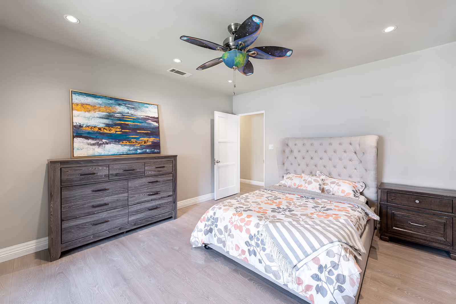 A bright bedroom with light gray walls and wood flooring. The room features a bed with a tufted headboard and floral bedding, a dark wooden dresser with a colorful abstract painting above it, and a matching nightstand. A ceiling fan with blades painted to resemble a galaxy hangs from the ceiling. The door to the room is open, showing a glimpse of a hallway.